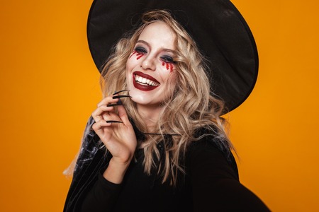 Portrait of blonde scary woman witch in black hat looking camera and smiling isolated over orange