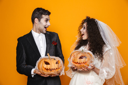 Photo of young zombie couple bridegroom and bride wearing wedding outfit and halloween makeup holding carved pumpkin isolated over yellow backgroundの写真素材