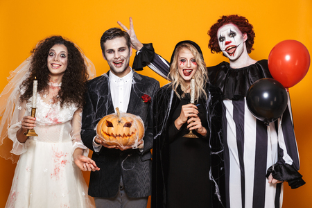 Group of cheerful dressed in scary costumes celebrating Halloween isolated over yellow background, holding balloons, curved pumpkinの写真素材