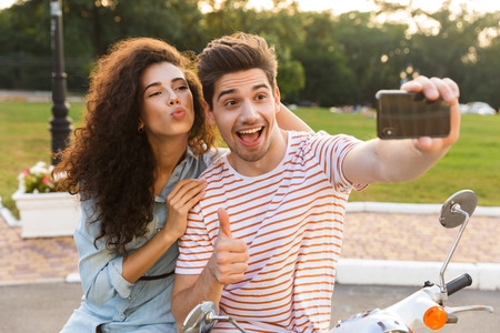 Photo of european couple man and woman taking selfie on mobile phone while sitting together on motorbike in city parkの写真素材