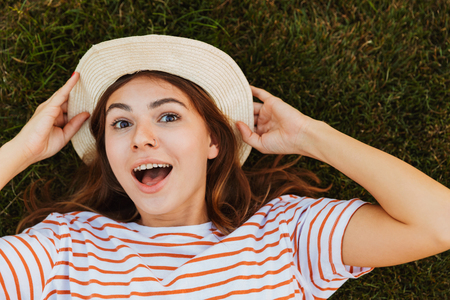 Top view of an excited young girl in summer hat laying on a grass, grimacingの写真素材