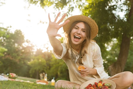 Smiling young girl in summer hat having a picnic at the park, sitting on a grass, outsretched handの写真素材