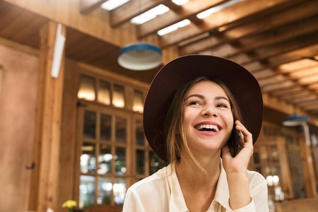 Close up of a laughing girl in hat sitting at the cafe table indoors, talking on mobile phoneの写真素材