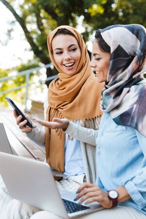 Photo of two muslim women wearing head scarfs using laptops and smartphone while sitting in parkの写真素材
