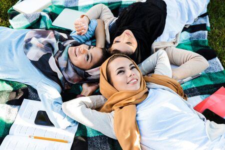 Photo from above of beautiful islamic women wearing headscarfs lying on blanket in green park while...