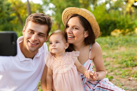 Pretty young family with little baby girl spending time together at the park, having a picnic, taking a selfieの写真素材