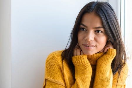 Close up portrait of a lovely young woman dressed in sweater sitting at the window indoors, looking awayの写真素材