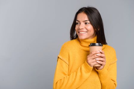 Portrait of a smiling young woman dressed in sweater holding takeaway coffee cup isolated over grey...