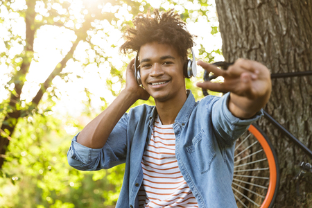 Cheerful young african teenager with bicycle outdoors, sitting on grass, listening to music with headphonesの写真素材
