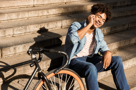 Image of cheerful young african guy sitting outdoors with bicycle on the street on steps talking by phone.の写真素材