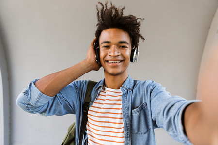 Image of cheerful young african guy standing outdoors listening music with headphones make selfie by camera.の写真素材