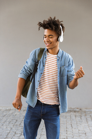 Cheerful young african man with backpack outdoors, listening to music with headphones, dancingの写真素材