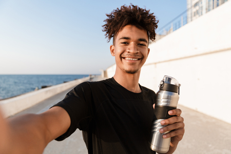Smiling young african teenager sportsman standing at the beach, taking a selfie with mobile phone, holding bottle of waterの写真素材