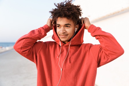 Close up of a handsome young african teenager dressed in hoodie standing at the beach, listening to music with earphonesの写真素材