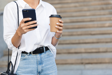 Cropped photo of young amazing woman on the street using mobile phone holding coffee.の写真素材