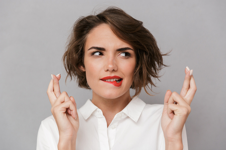Portrait of a worried young woman standing isolated over gray background, holding fingers crossed for good luckの写真素材