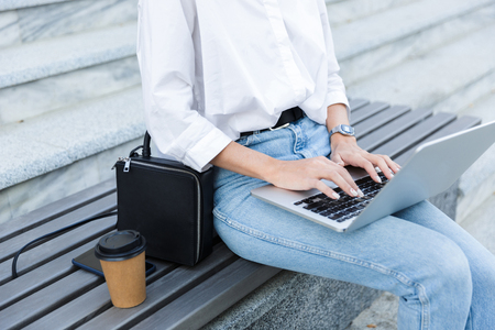 Close up of a young woman sitting on bench outdoors at the street, using laptop computerの写真素材