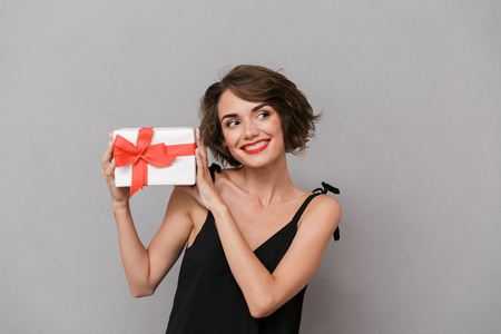 Photo of adorable woman 20s wearing black dress holding present box isolated over gray backgroundの写真素材