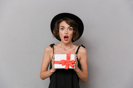 Photo of surprised woman 20s wearing black dress and hat holding present box isolated over gray backgroundの写真素材