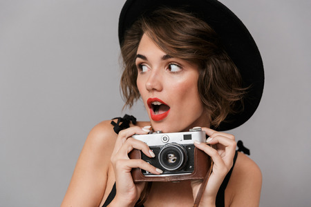 Photo of positive woman 20s wearing black dress and hat holding retro camera isolated over gray...