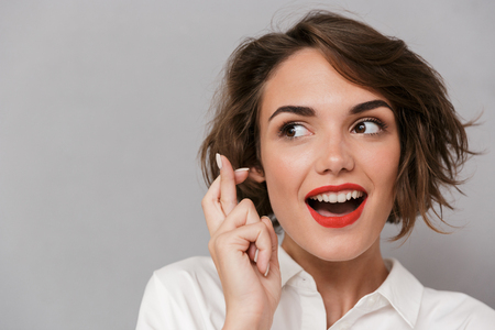 Portrait of a worried young woman standing isolated over gray background, holding fingers crossed for good luck, looking awayの写真素材