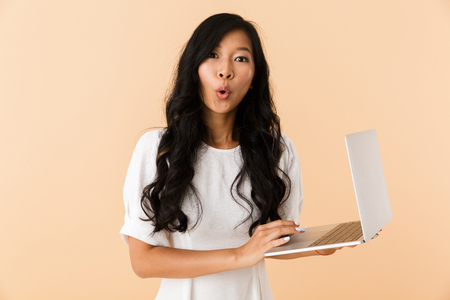 Portrait of an excited young asian woman isolated over beige background, standing , using laptopの写真素材
