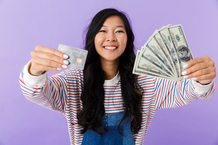 Portrait of a happy young asian woman isolated over violet background, holding plastic credit card, showing money banknotesの写真素材
