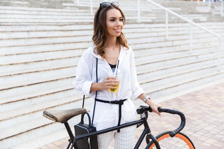 Happy young woman spending time at the park, walking with bicycle, drinking lemonadeの写真素材