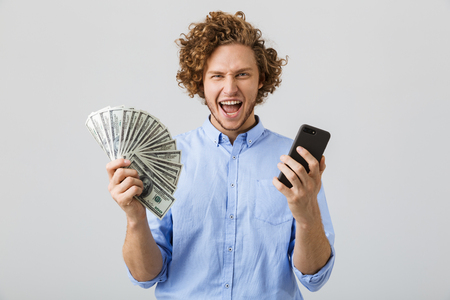Portrait of a positive young man with curly hair isolated over white background, showing money banknotes, holding mobile phoneの写真素材