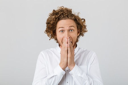 Portrait of a surprised young man with curly hair isolated over white background, cover mouth with...