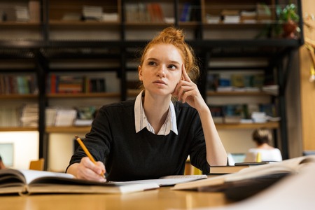 Concentrated red haired teenage girl studying at the table in library
