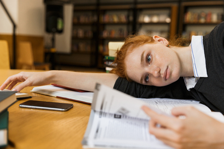 Tired red haired teenage girl studying at the table in library, laying on a tableの写真素材