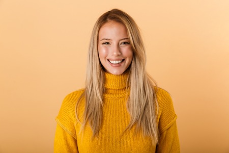 Portrait of a smiling young woman dressed in sweater standing isolated over yellow background, looking at cameraの写真素材