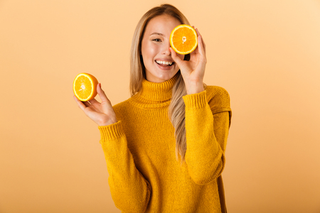 Portrait of a happy young woman dressed in sweater standing isolated over yellow background, holding sliced orange at her faceの写真素材