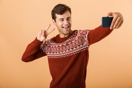 Portrait a smiling young man standing isolated over beige background, taking selfie with mobile phoneの写真素材