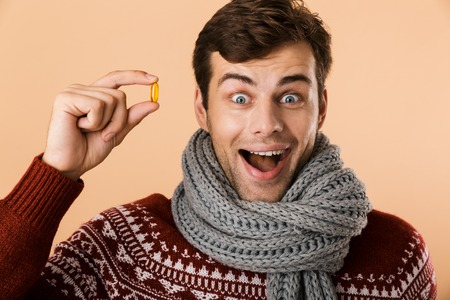 Close up portrait an excited man dressed in sweater and scarf isolated over beige background, showing pills capsulesの写真素材