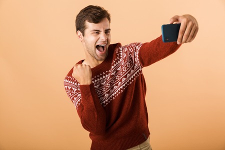 Portrait a smiling young man standing isolated over beige background, taking selfie with mobile phone, celebratingの写真素材