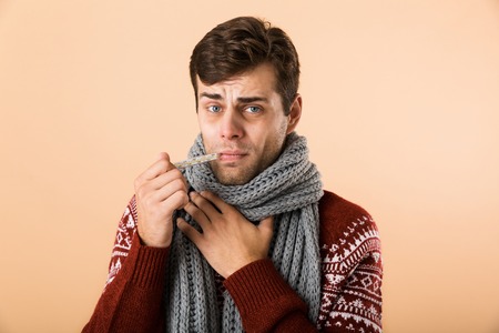 Portrait of a sick young man dressed in sweater and scarf isolated over beige background, holding thermometer in his mouthの写真素材
