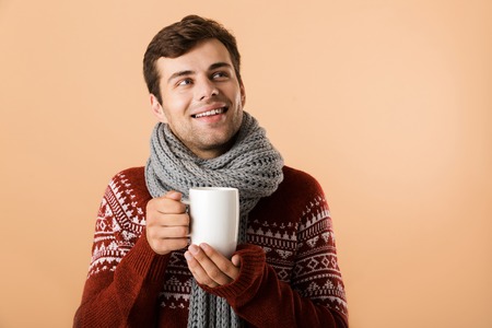 Close up of a smiling young man dressed in sweater and scarf drinking tea from a cup isolated over beige backgroundの写真素材