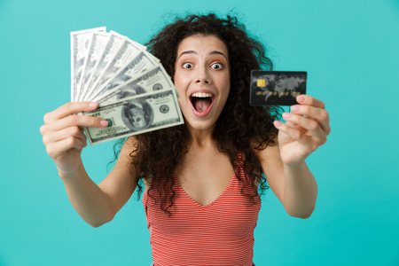 Image of delighted woman 20s wearing casual clothing holding fan of dollar money and credit card standing isolated over blue backgroundの写真素材