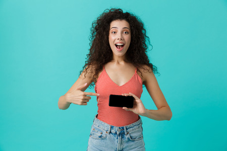 Photo of charming woman 20s with curly hair smiling and holding smartphone isolated over blue backgroundの写真素材