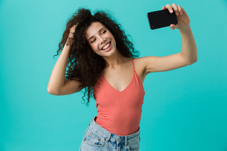 Photo of young woman 20s with curly hair taking selfie on mobile phone isolated over blue background