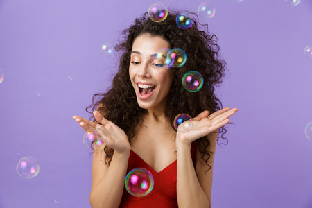 Image of young woman 20s wearing red dress laughing and standing under falling soap bubbles isolated...