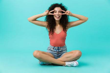 Image of charming woman 20s wearing casual clothing laughing while sitting on floor with legs crossed isolated over blue backgroundの写真素材