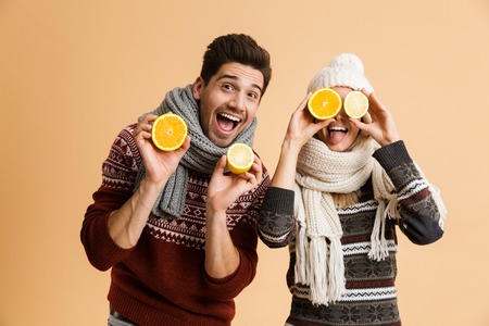 Portrait of a smiling young couple dressed in sweaters and scarves standing together isolated over beige background, showing sliced lemons and orangesの写真素材