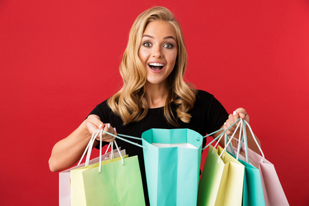 Portrait of charming blond woman 20s smiling and holding colorful shopping bags isolated over red background in studioの写真素材