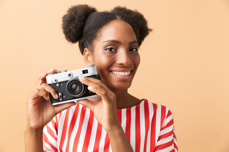 Happy african american woman smiling and photographing on retro camera isolated over beige backgroundの写真素材