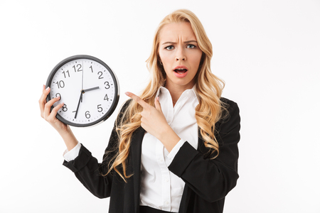 Portrait of a shocked young businesswoman standing isolated over white background, showing clock, pointing fingerの写真素材
