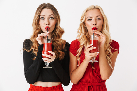 Portrait of two cheerful young smartly dressed women wearing makeup standing isolated over white background, celebrating, drinking tomato juice from glassesの写真素材