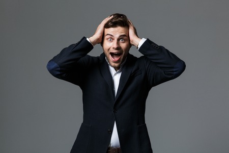 Portrait of a shocked young businessman dressed in suit isolated over gray background, celebrating...